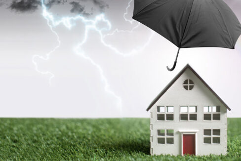 Haus auf einer grünen Wiese, dass vor Wetterergnissen und anderen Schadensfällen durch einen schwarzen Regenschirm geschützt wird.